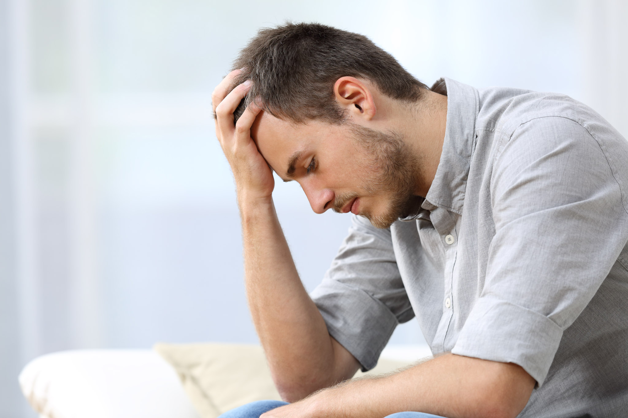 Side view of a sad man with a hand on the head sitting on a couch in the living room at home